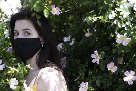 Attractive young brunette woman in yellow t-shirt and medical black face mask. in bush with pink...