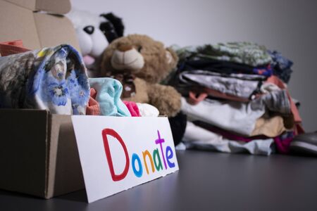 Donate sign handwritten with multicoloured letters. A box with clothes and a pile of clothes nearby on a grey table.clothes donation concept. copy space. High quality photoの写真素材