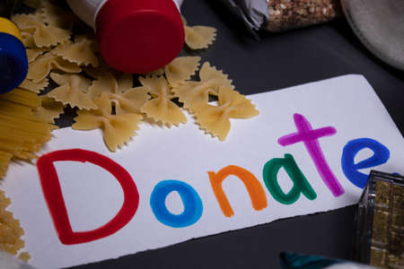 Donate sign handwritten with multicoloured letters. A pile of food supplies on a grey table. Food donation concept. Context of The Coronavirus Pandemic. copy space. High quality photoの写真素材