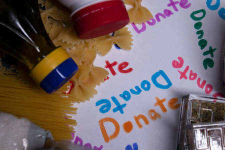 Donate sign handwritten with multicoloured letters. A pile of food supplies on a grey table. Food donation concept. Context of The Coronavirus Pandemic. copy space. High quality photoの写真素材