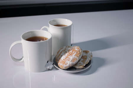 two white cups with tea and sticking out tea bags next to gingerbread cookies heart-shaped on a white table in a gray kitchen. High quality photoの写真素材