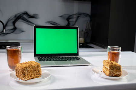 two cups of tea and two pieces of cake in gray kitchen next to laptop with green screen - chromakey....