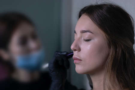 the process of applying makeup to a beautiful brunette by a master in a medical mask and black...