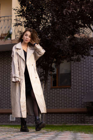 attractive slim young brunette in fashionable trench coat and black dress walking towards. autumn, yellow leaves. brick building background.の写真素材