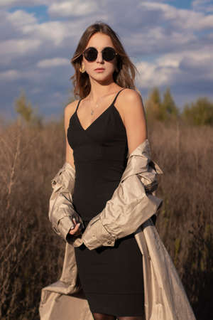 brunette in fashionable trench coat, black dress and sunglasses in the field. autumn, yellow leaves, blue sky.の写真素材
