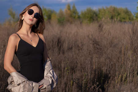 brunette in fashionable trench coat, black dress and sunglasses in the field. autumn, yellow leaves, blue sky.の写真素材