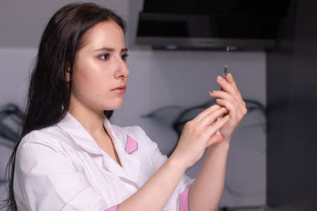 attractive brunette nurse in white and pink medical gown with a syringe in gray room. healthcare,...