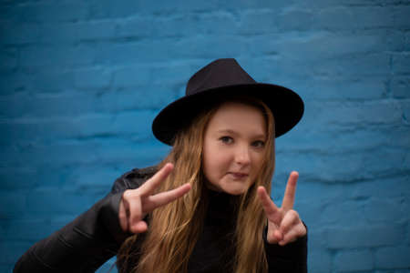 portrait of cute brunette teen girl in Leather Jacket and black hat showing peace sign, on the...