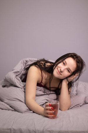 charming sleepy brunette woman with glass cup of tea in bed with grey bed linens. morning routine....