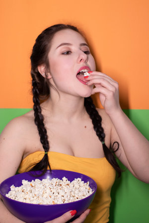 charming brunette woman in yellow top holding a purple bowl with popcorn on bright green and orange...