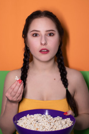 charming brunette woman in yellow top holding a purple bowl with popcorn on bright green and orange background. movie concept. cinema themeの写真素材