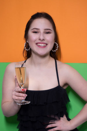 charming brunette woman in black dress holding glass of champagne with an engagement ring inside on bright green and orange background. wedding concept. engagement. poping the question. brideの写真素材