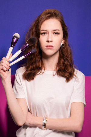 charming brunette woman in white t-shirt with makeup brushes isolated on colorful blue and pink background. professional makeup artist. copy spaceの写真素材