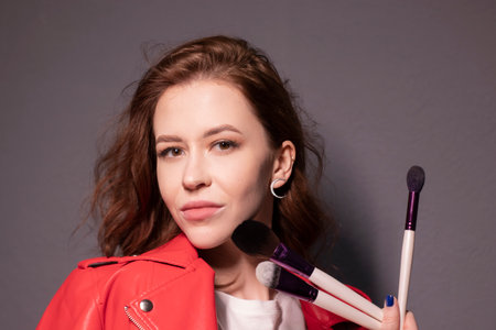 charming brunette woman in red leather jacket with makeup brushes isolated on grey wall background. professional makeup artist. copy spaceの写真素材