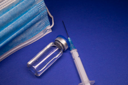 Vaccine bottle phial with no label near blue medical mask and medical syringe with injection needle. isolated on blue background. cure.の写真素材