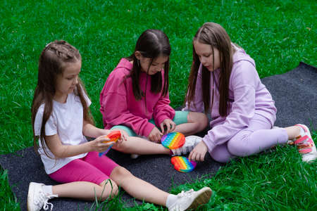 three teenage girls in colorful clothes play with trendy sensory toy pop it outdors. rainbow soft silicone bubbles. anxiety relif, antistress, mental health concept. Anti stress game popit.の写真素材
