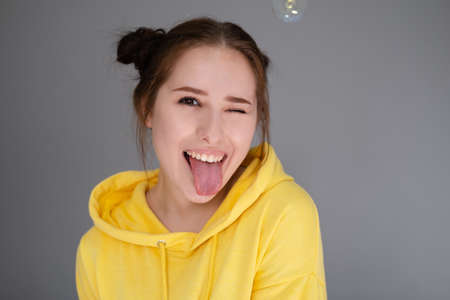 happy brunette woman in yellow hoodie among soap bubbles in bright gray room. happy people. millennial generation. fashionable teenager. trendy colorsの写真素材