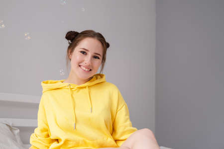 cheerful positive brunette woman in yellow hoodie in white bedroom on bed with white linens. happy...