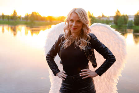 sexy blonde woman in black leather jacket and shorts with white angel wings. demon or angel in hell or heaven. sunset near lake.の写真素材