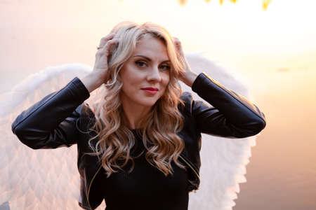 sexy blonde woman in black leather jacket and shorts with white angel wings. demon or angel in hell or heaven. sunset near lake.の写真素材