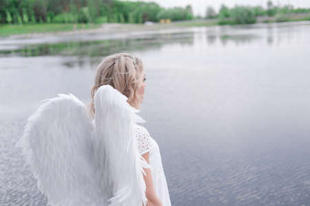 charming blonde with white angel wings standing by the water. beautiful woman in angel costume. goddess.の写真素材