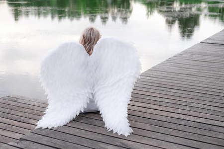 charming blonde with white angel wings standing by the water. beautiful woman in angel costume. goddess.の写真素材