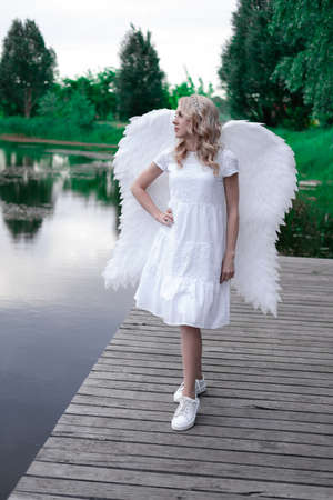 charming blonde with white angel wings standing by the water. beautiful woman in angel costume. goddess.の写真素材