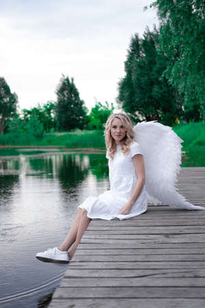 beautiful angel in white dress. Amazing blond woman with long curly hair and white wingsの写真素材