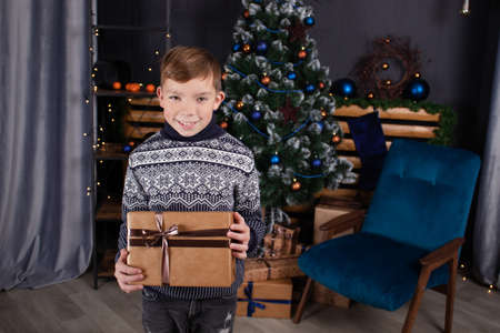 happy handsome pretty teenage blonde boy in knitted sweater holding new years present near a christmas tree.の写真素材