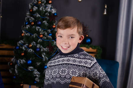happy handsome pretty teenage blonde boy in knitted sweater holding new years present near a christmas tree.の写真素材