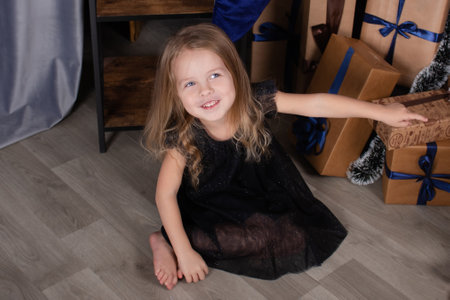 charming preschool blonde girl in a black dress standing near a christmas tree with blue decorations in dark room.の写真素材