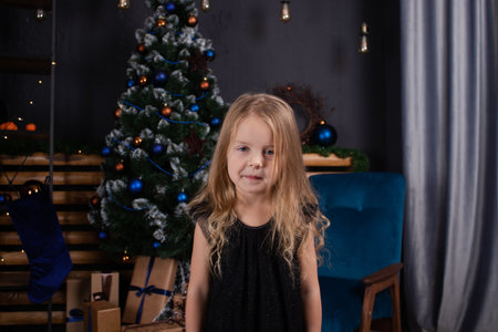 charming preschool blonde girl in a black dress standing near a christmas tree with blue decorations in dark room.の写真素材