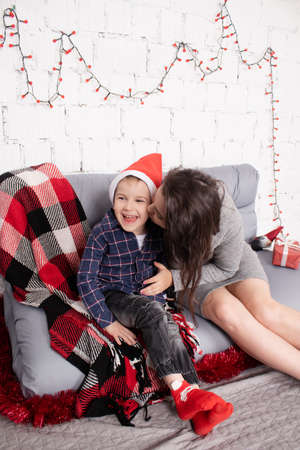 happy incomplete family - mother and son - hugging on grey couch covered in red checkered plaid-blanket near red christmas decor. cozy young family. single mother.の写真素材