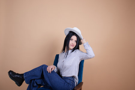 portrait of young woman in white blouse, white hat and jeans near blue chair on brown background. pretty female portrait.の写真素材