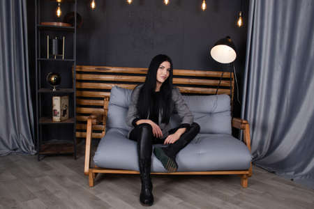 confident brunette woman in black in grey loft room sitting on a couch. trendy clothes. fashion...