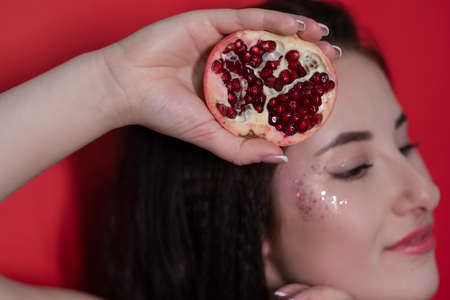 charming brunette woman girl female with cropped half pomegranate near skin face on red background. tasty juicy fruit.の写真素材