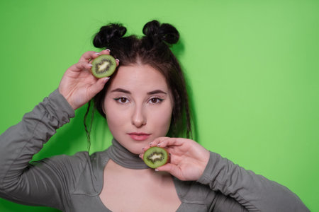 Beauty portrait of attractive young woman with clean radiant skin face with kiwi halves, brunette girl with bare shoulders holding kiwi on green background. Tropical fruits, Healthy food.の写真素材