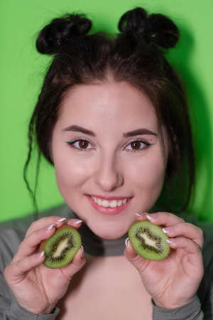 young woman with kiwi halves, brunette girl with bare shoulders holding kiwi near face on green background. Tropical fruits, Healthy food. tasty juicy fruitの写真素材