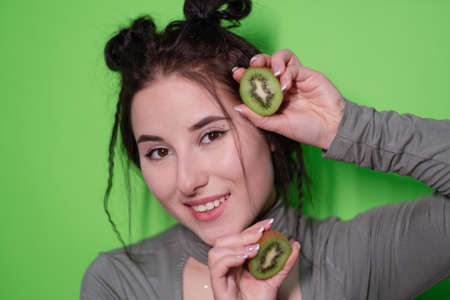 Beauty portrait of attractive young woman with clean radiant skin face with kiwi halves, brunette girl with bare shoulders holding kiwi on green background. Tropical fruits, Healthy food.の写真素材