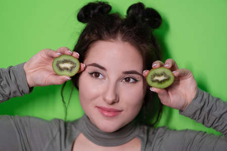 young woman with kiwi halves, brunette girl with bare shoulders holding kiwi near face on green background. Tropical fruits, Healthy food. tasty juicy fruitの写真素材