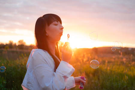 charming brunette woman making inflating soap bubbles in field on sunset.