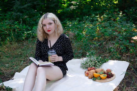 pretty blonde girl on a picnic in a forest or park with lemonade, fruits and croissants. summertime, rest, relaxation. millennial generation.の写真素材