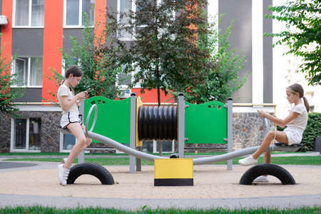 two teenage girls using smartphones outside near black and yellow warning or attention sign with...