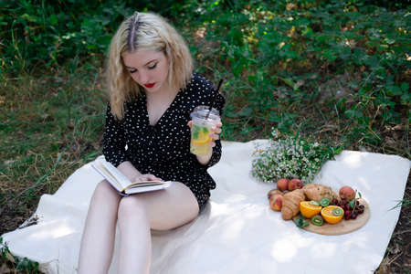 portrait of young woman on a picnic on plaid in park reading a book with tasty snacks.の写真素材