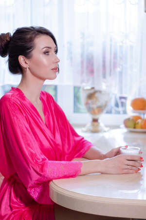 young brunette woman in kitchen wearing pink dressing gown with a cup of hot beverage coffee. tender woman portrait during morning routine and breakfast.の写真素材