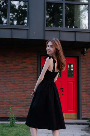 charming young woman in a black dress in front of modern building with red door. housewarming. buying a house happy life in private house.の写真素材