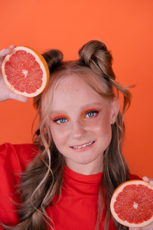young girl with grapefruit slice on orange background. curly hair and two ponytails.の写真素材