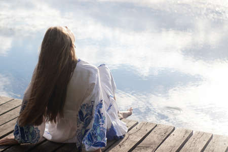 a girl in an embroidered Ukrainian shirt sits on the pier, the reflection of clouds in the water of the lake. On the shore of the sky. vyshyvanka day. freedom. patriot.の写真素材