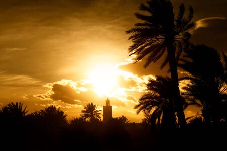 Minaret of a mosque at sunset with moddy orange sky and dark palms. Concept of islam, prayer, religion and faith.の写真素材