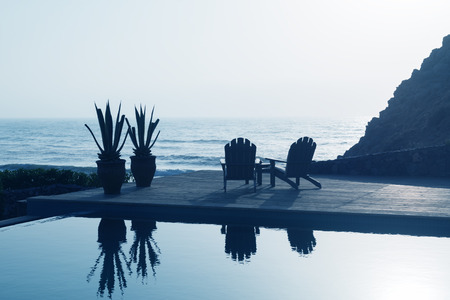 Two wooden relaxing chairs next to a swimming pool overlooking the sea.